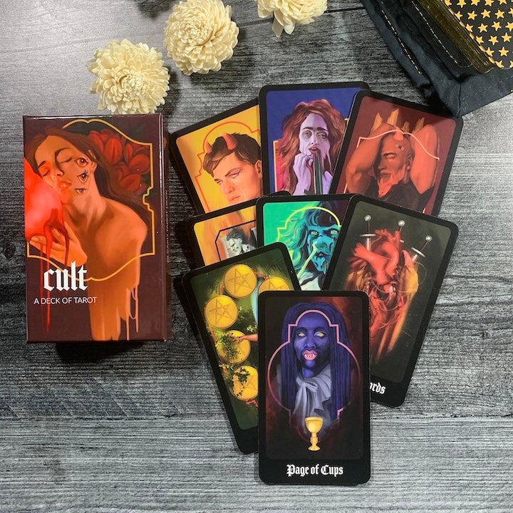 Cult Tarot Deck with a shiny burgundy box and a figure of a woman sensually hugging a glowing red heart. she has three eyes on the side of her head. There are 8 different tarot cards splayed out next to the box with different creepy images across them including a vampire with purple skin.